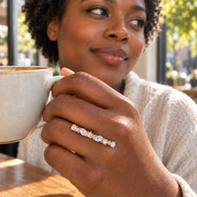 A yellow gold gemstone ring is worn on a hand holding a speckled ceramic mug beside a sunlit cafe window.