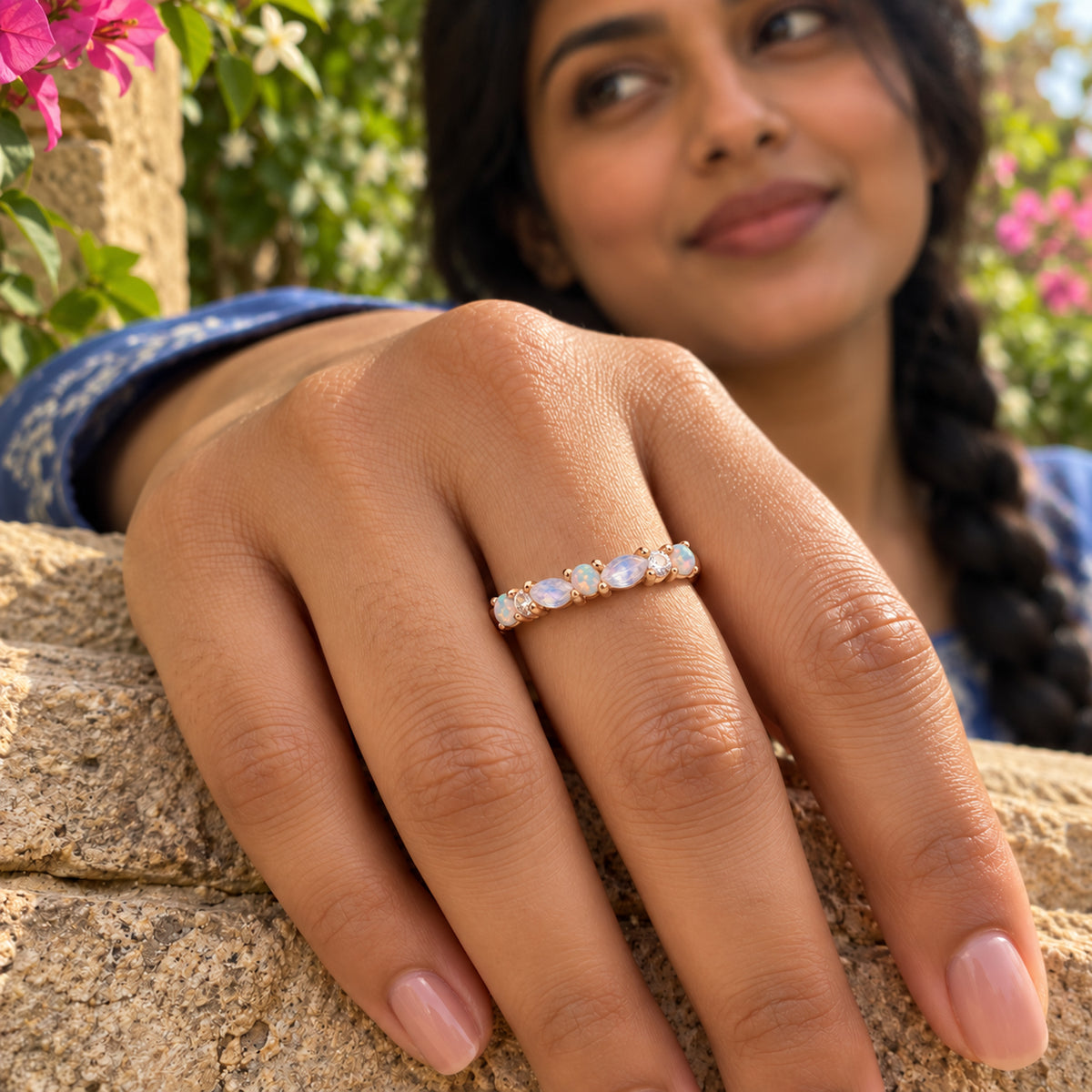A rose gold gemstone ring is worn on a hand resting on a stone wall with pink flowers in the background.