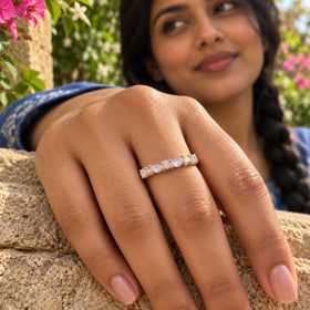 A rose gold gemstone ring is worn on a hand resting on a stone wall with pink flowers in the background.