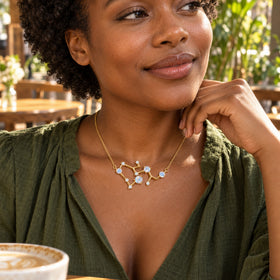 A woman with short curly hair wears a yellow gold necklace with an angular stone pendant above an olive blouse in a sunlit cafe.