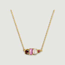 A yellow gold vermeil necklace displays a burgundy round gem, a rectangular pink gem, a white pearl and a small pink round gem on a pale gray background.