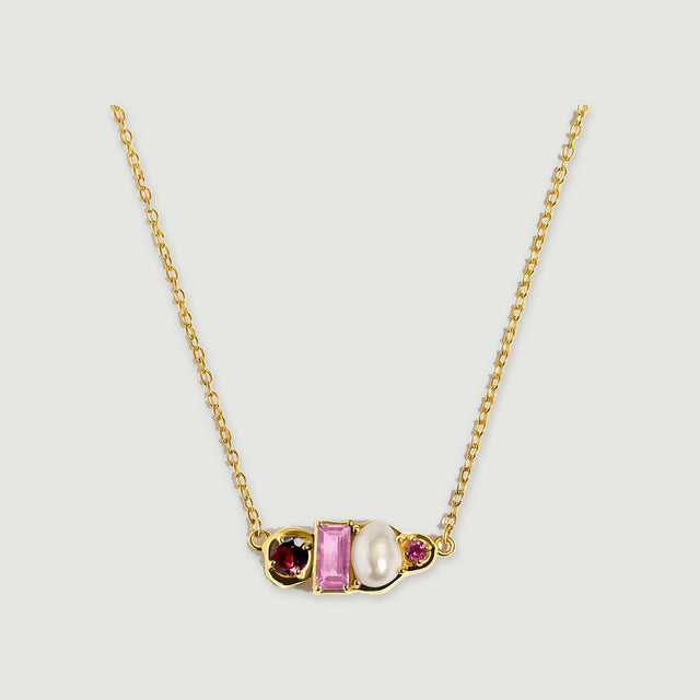 A yellow gold vermeil necklace displays a burgundy round gem, a rectangular pink gem, a white pearl and a small pink round gem on a pale gray background.