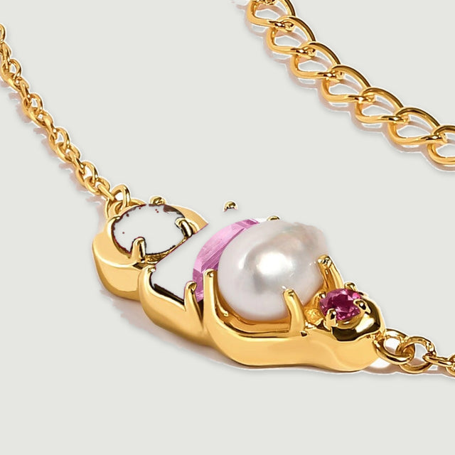 An angled yellow gold necklace pendant shows the raised pearl, pink rectangular stone and burgundy round stone beside the chain.