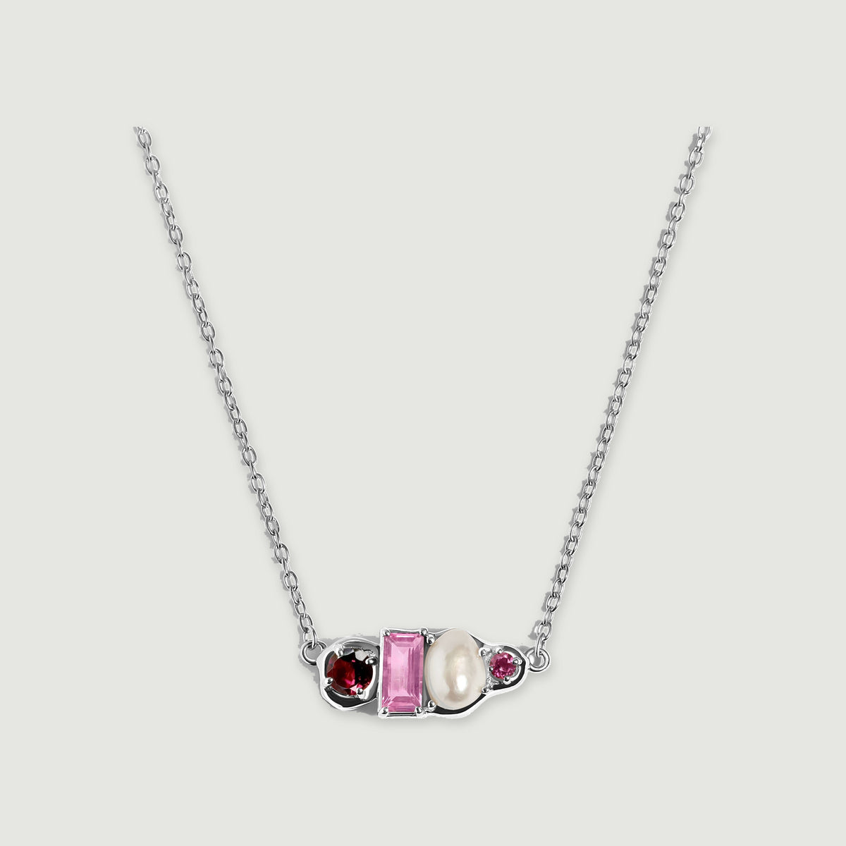 A sterling silver chain curves toward an asymmetric pendant with burgundy and pink stones beside a white pearl.