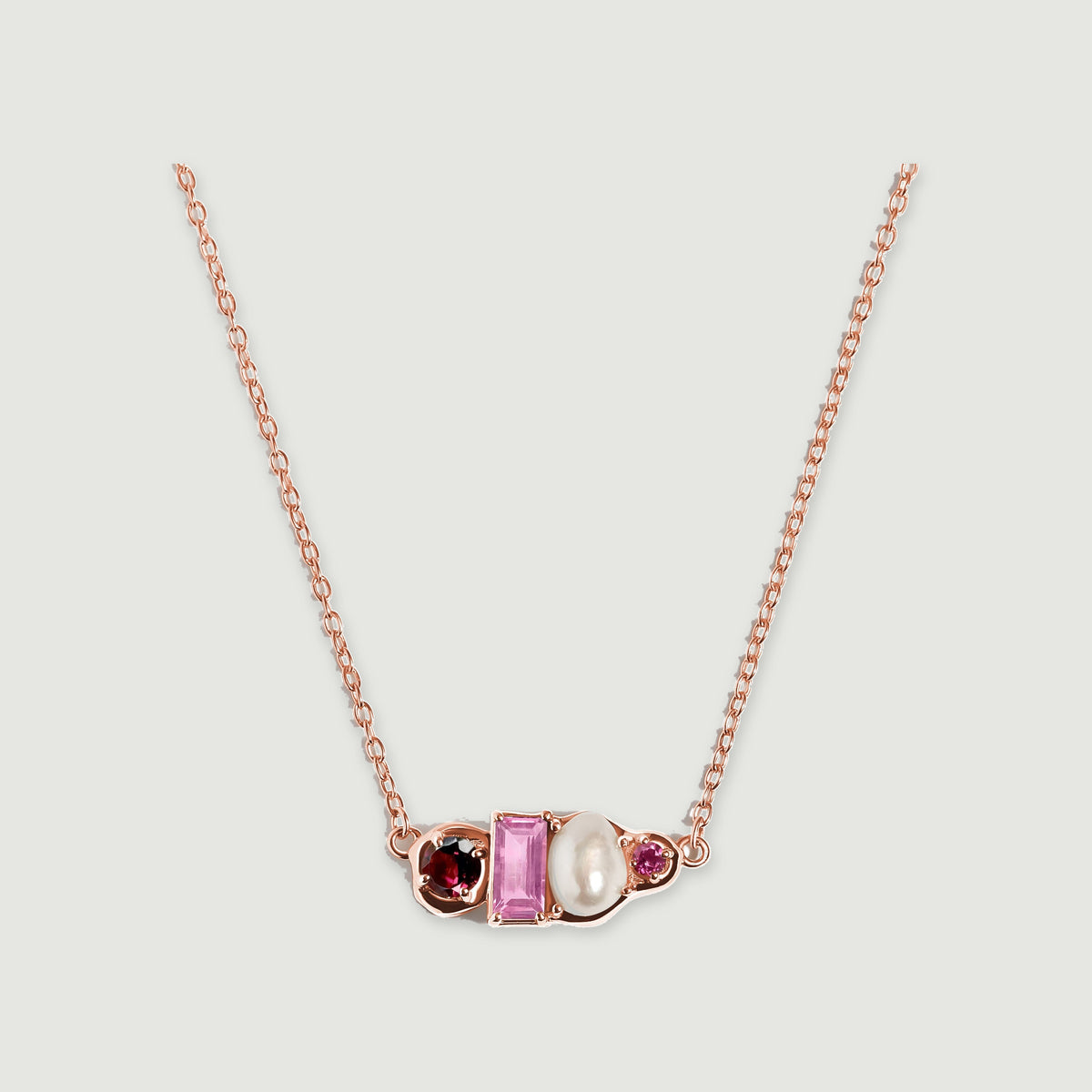 A rose gold vermeil necklace rests against a pale gray surface with its four differently shaped pendant elements facing forward.