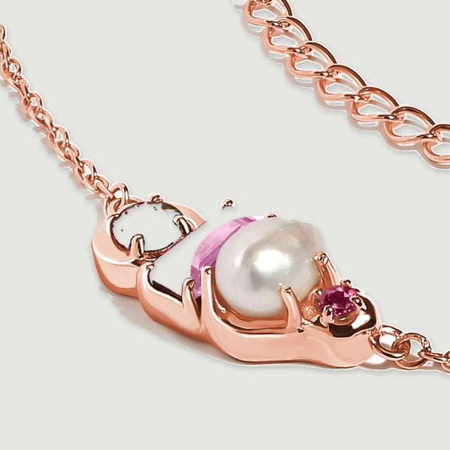 A rose gold necklace close-up shows side prongs around a white pearl and pink stones with burgundy garnet on the left.