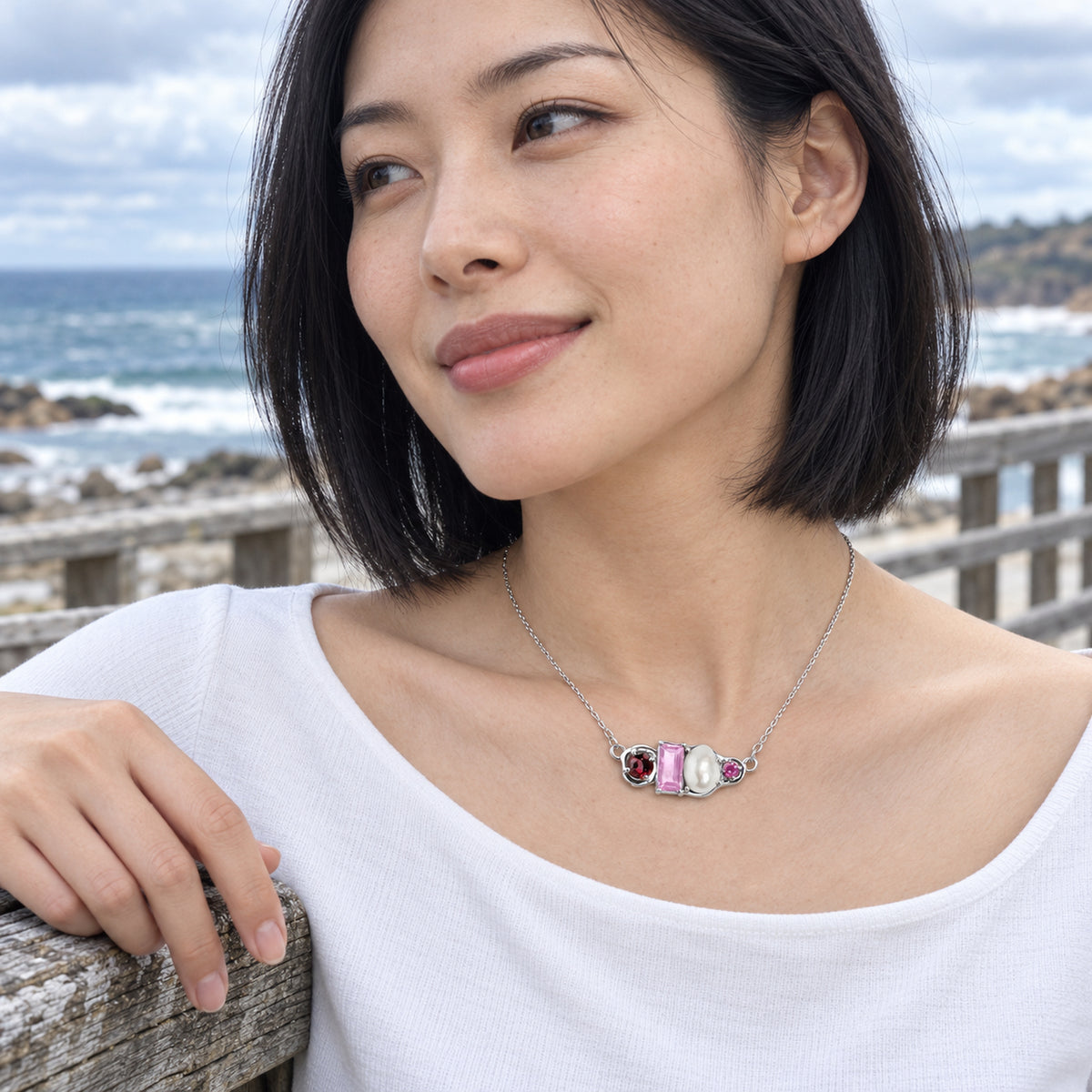 A woman with short black curls wears a silver chain and a four-part gemstone and pearl pendant above an open white shirt in a leafy garden.