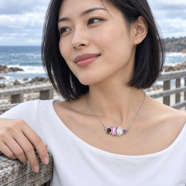 A woman with short black curls wears a silver chain and a four-part gemstone and pearl pendant above an open white shirt in a leafy garden.