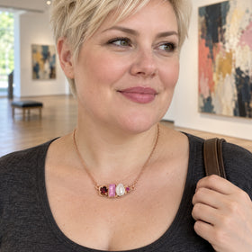 A blonde woman in a sage green cardigan wears a rose gold necklace with a horizontal pink and burgundy gemstone pendant beside a white pearl.