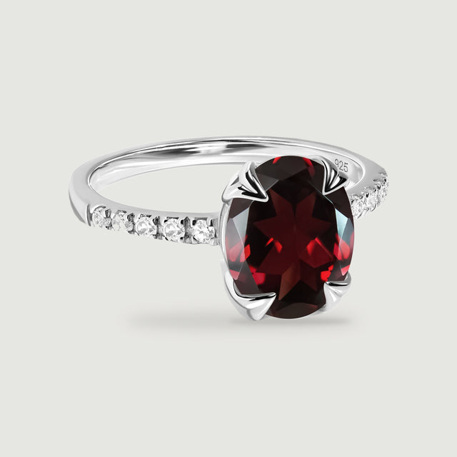 A polished silver ring frames a faceted red oval stone between two rows of clear accents.