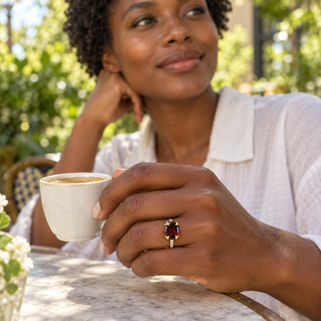 A yellow gold garnet ring is worn on a hand holding a ceramic coffee cup at an outdoor table.