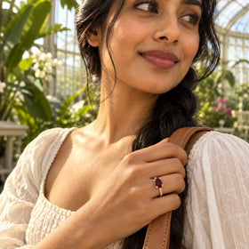 A rose gold garnet ring is worn on a hand gripping a tan bag strap among sunlit plants.