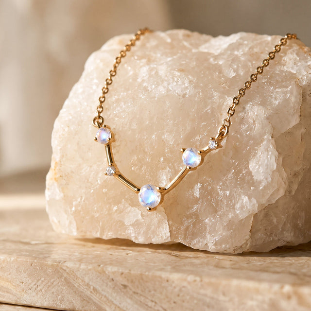 The gold necklace is draped across a pale textured stone on a clean beige tabletop.