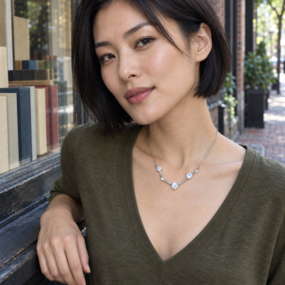A woman with a short black bob wears the silver necklace with an olive V-neck top beside a bookshop window.