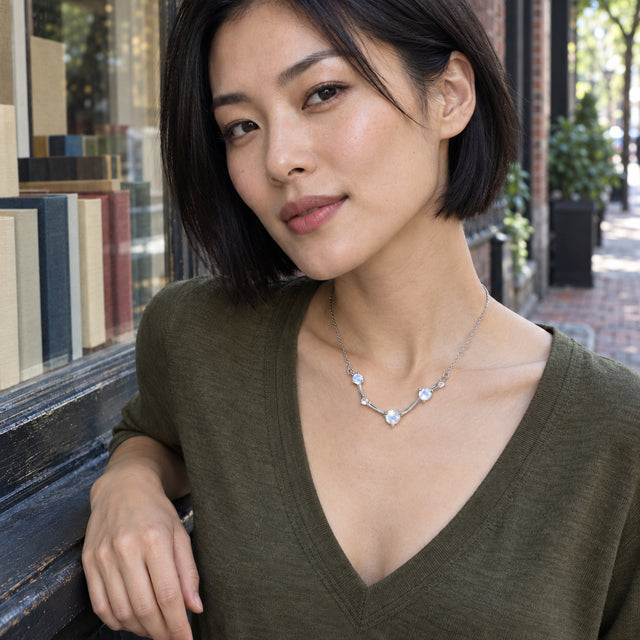 A woman with a short black bob wears the silver necklace with an olive V-neck top beside a bookshop window.