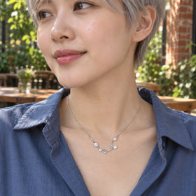 A small silver moonstone necklace rests above a blue shirt with sunlit cafe greenery and brick behind the wearer.