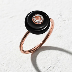 The rose gold vermeil ring rests on a light stone surface, showing its black disc, center stones, and rounded band.