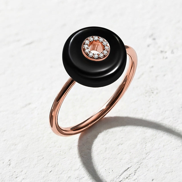 The rose gold vermeil ring rests on a light stone surface, showing its black disc, center stones, and rounded band.