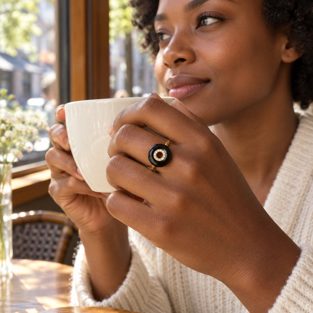 A yellow gold vermeil ring with a black disc and clear center stones is worn on a hand holding a white cup beside a cafe window.