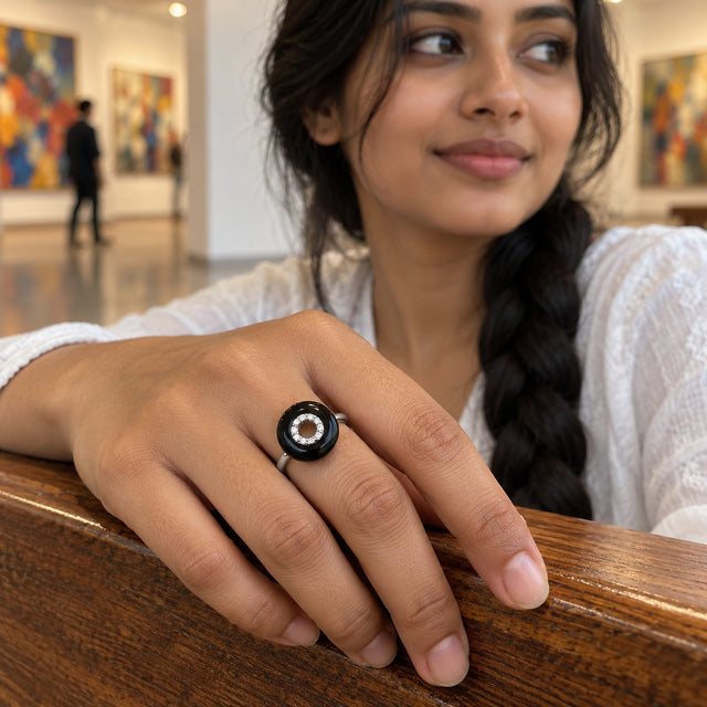 A sterling silver ring with a glossy black disc and clear center stones is worn on a hand resting on a wooden gallery bench.