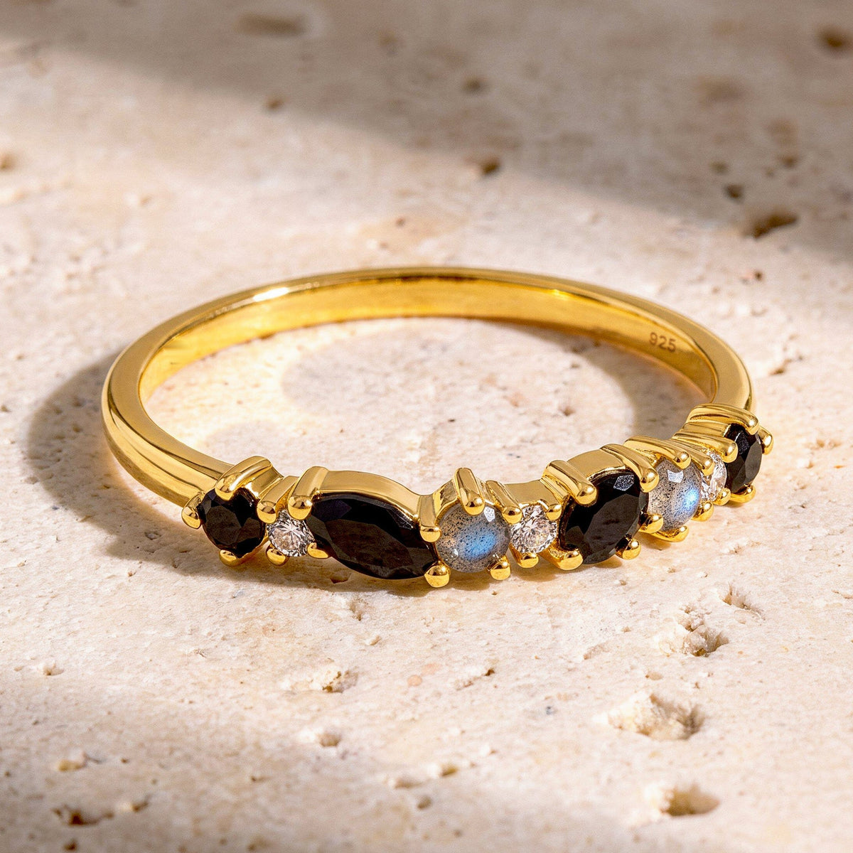 The yellow gold ring lies on a pale stone surface with dark faceted gems and blue flashes in the round accent stones.