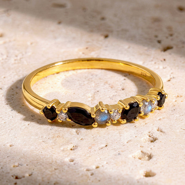 The yellow gold ring lies on a pale stone surface with dark faceted gems and blue flashes in the round accent stones.