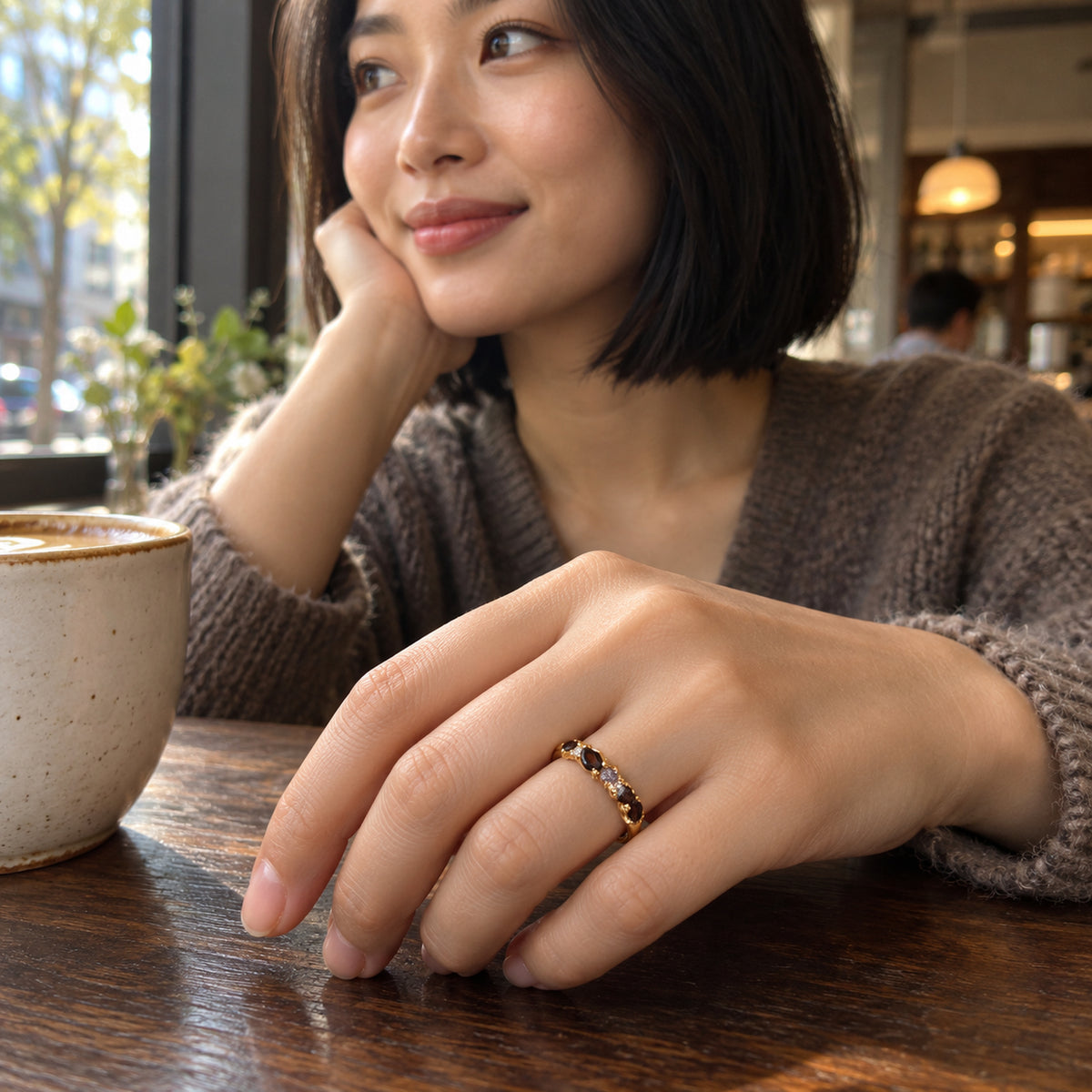 A woman wears a yellow gold ring with dark stones and clear accents while resting her hand beside a ceramic coffee cup in a cafe.