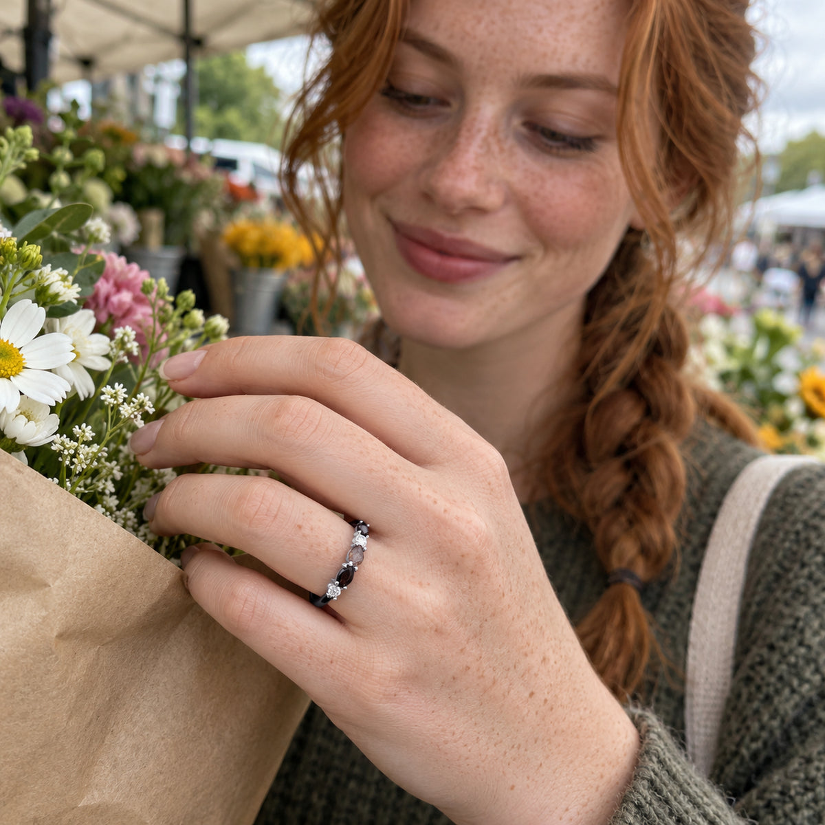 A freckled woman wears a silver ring with dark and clear stones as she holds a paper-wrapped bouquet at a flower stall.