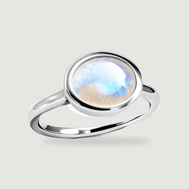 A polished sterling silver ring with an oval blue-flashing moonstone stands at an angle on a light gray surface.