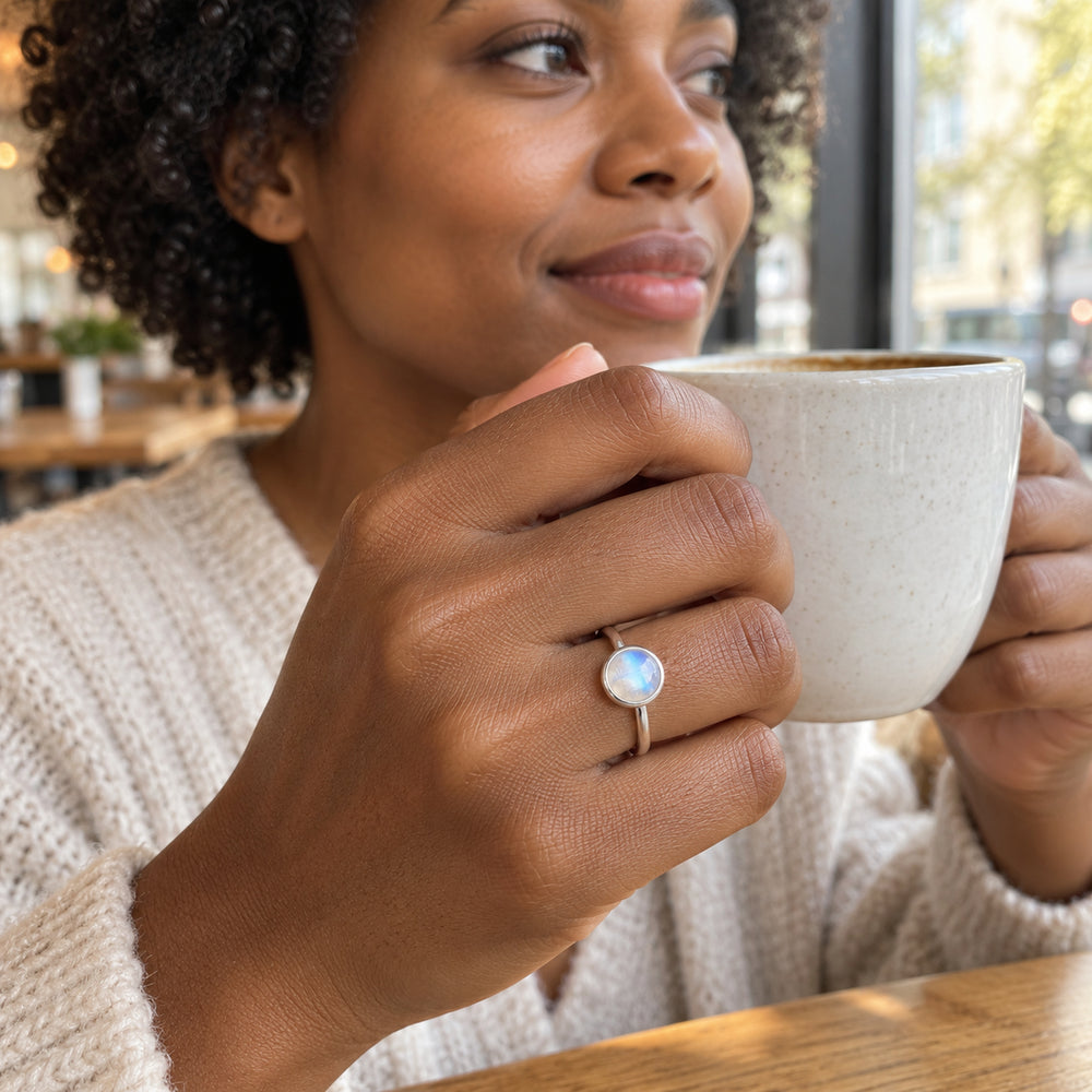 A woman wearing a silver moonstone ring holds a cream ceramic cup beside a cafe window.