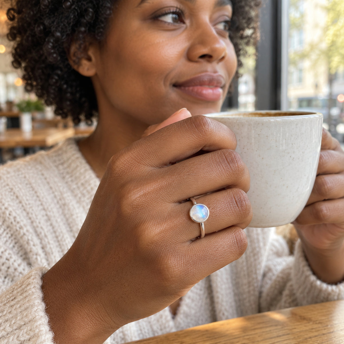 A woman wearing a silver moonstone ring holds a cream ceramic cup beside a cafe window.