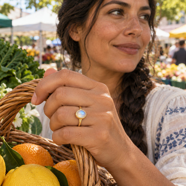 A yellow gold moonstone ring is visible on a hand holding a wicker basket filled with citrus at an outdoor market.