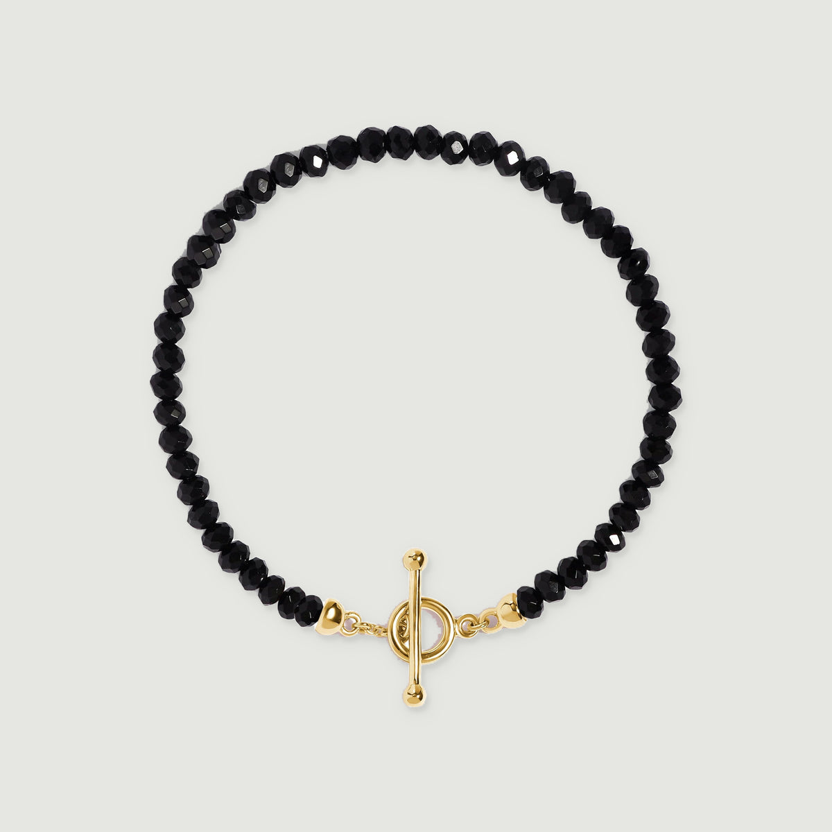 An oval strand of faceted black beads ends in a yellow gold vermeil toggle clasp against a pale gray surface.