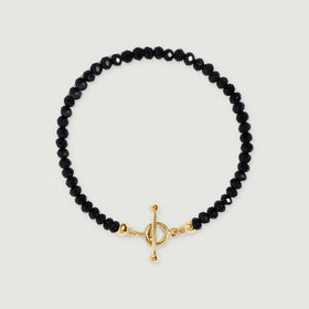 An oval strand of faceted black beads ends in a yellow gold vermeil toggle clasp against a pale gray surface.