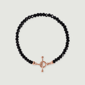 A black obsidian bead bracelet lies centered on light gray with a rose gold vermeil ring and slender bar closure.