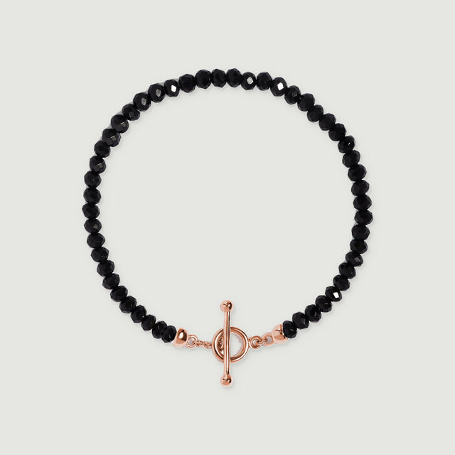 A black obsidian bead bracelet lies centered on light gray with a rose gold vermeil ring and slender bar closure.