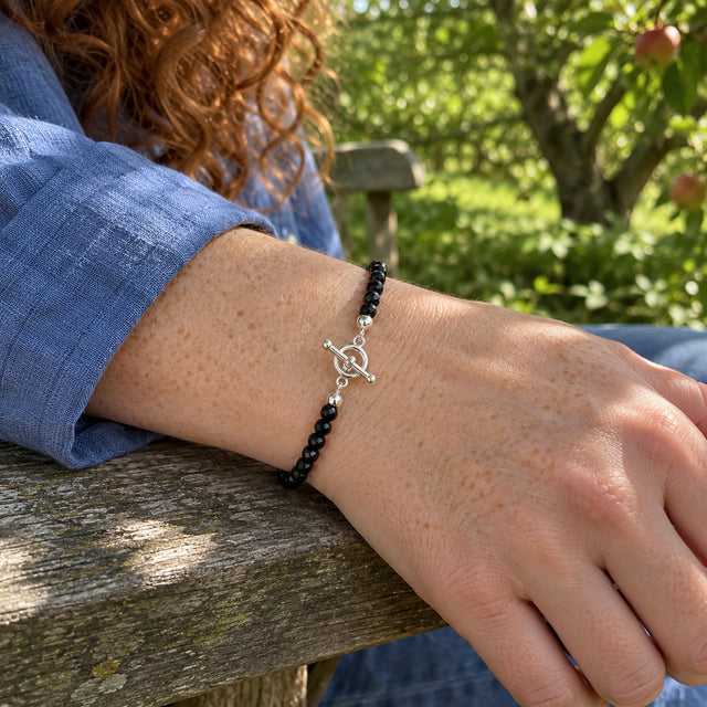 Faceted black obsidian beads and a silver ring-and-bar clasp circle a freckled wrist beside a blue linen sleeve on a weathered wooden bench.