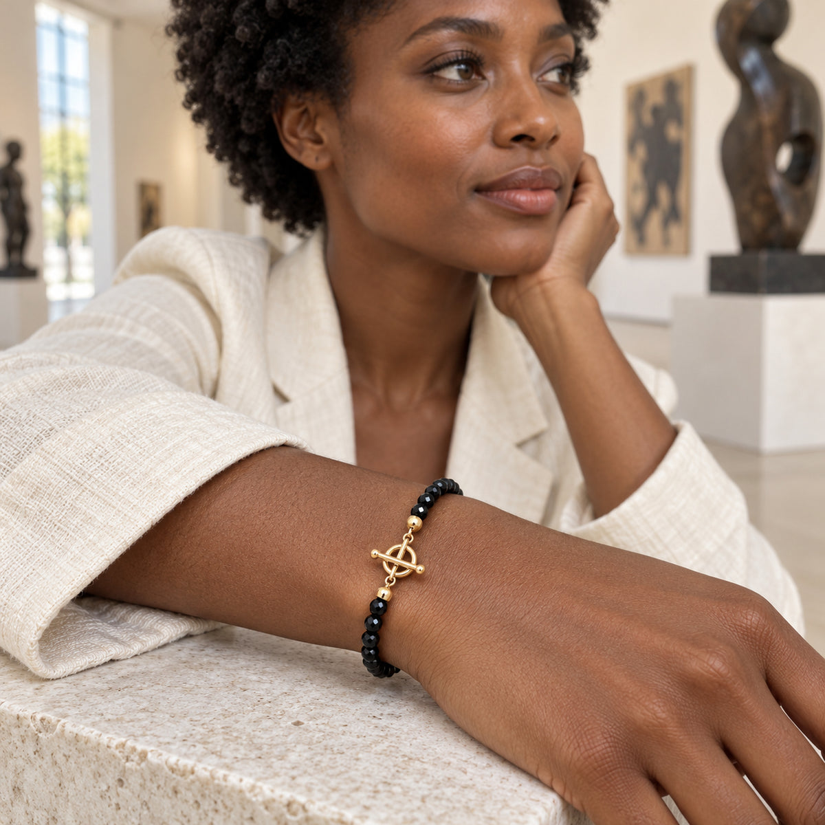 A yellow gold vermeil toggle clasp and black faceted beads encircle a woman's wrist resting on a pale stone ledge in a gallery.