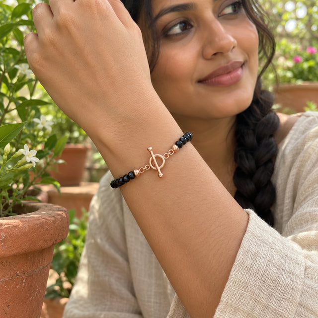 A rose gold vermeil ring-and-bar clasp joins black faceted beads around a raised wrist among green leaves and terracotta planters.