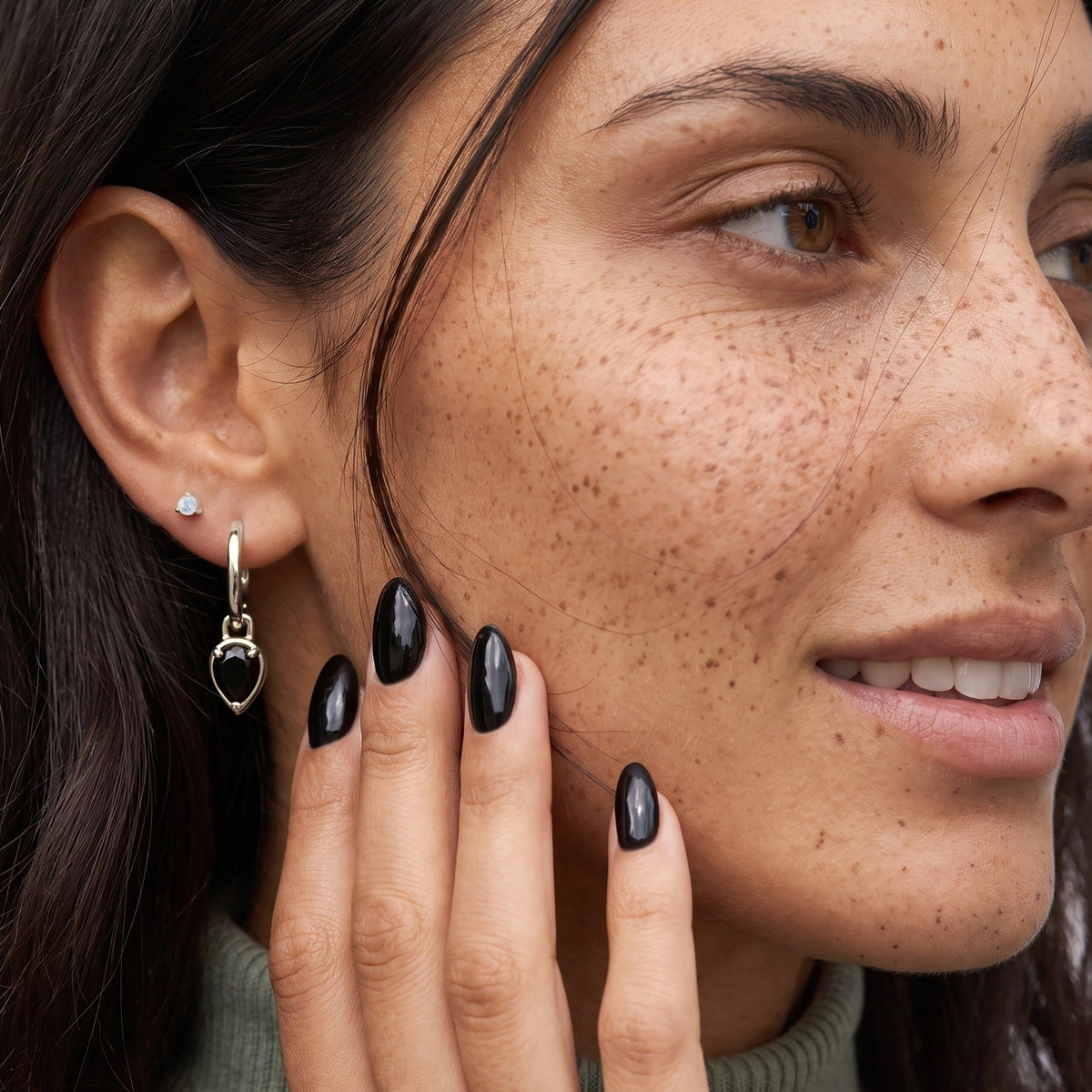 Freckled person touches a black onyx pear-drop hoop earring while wearing a small stud.