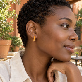 A yellow gold plated carnelian hoop is worn by a woman with short curls at a leafy outdoor cafe.
