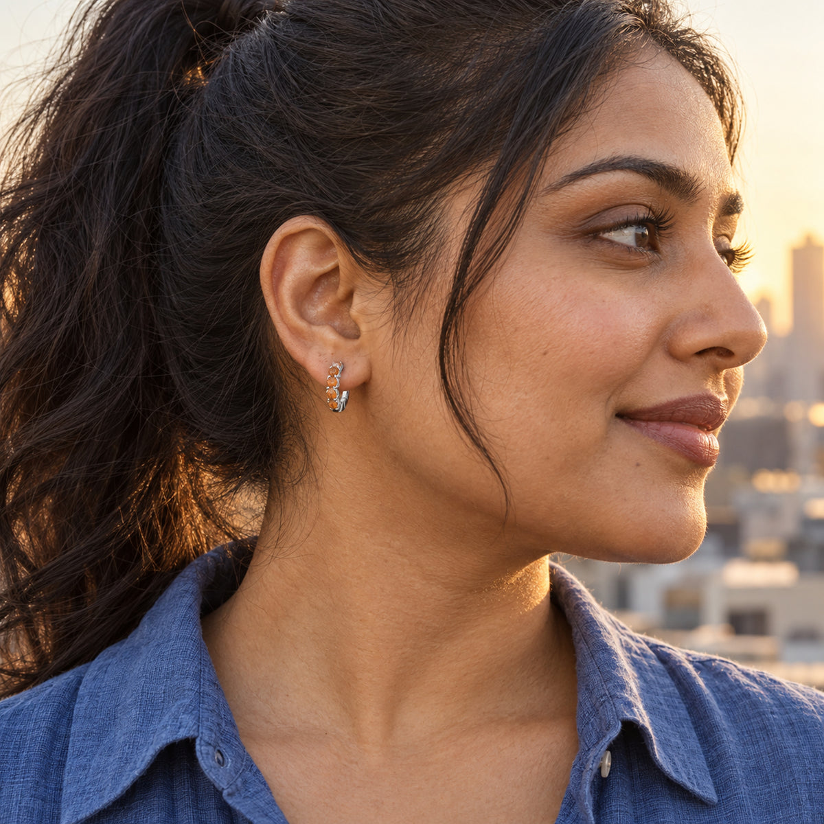 A woman with a dark ponytail wears a white rhodium plated carnelian hoop above a blue shirt with a city skyline behind her.