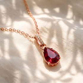 A rose gold ruby necklace lies across softly folded ivory fabric in dappled natural light.