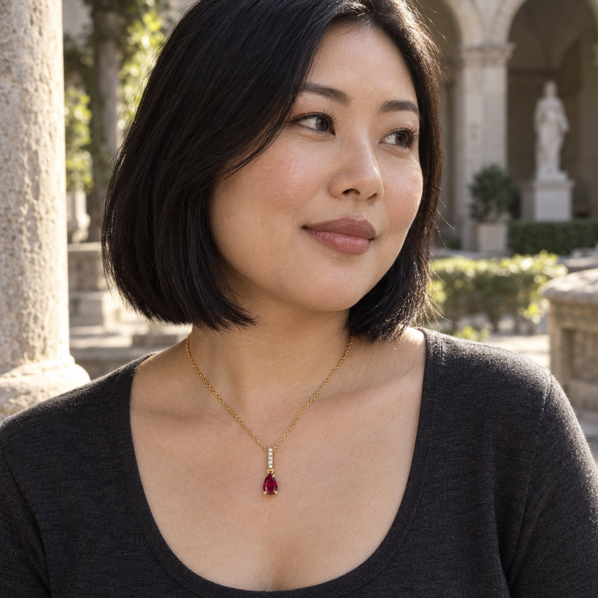 A woman in a charcoal scoop-neck top wears a yellow gold ruby necklace in a sunlit stone courtyard.