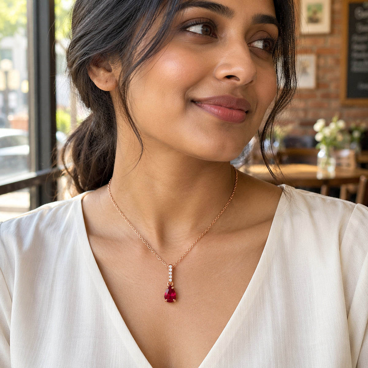 A woman in an ivory V-neck blouse wears a rose gold ruby pendant beside a bright cafe window.