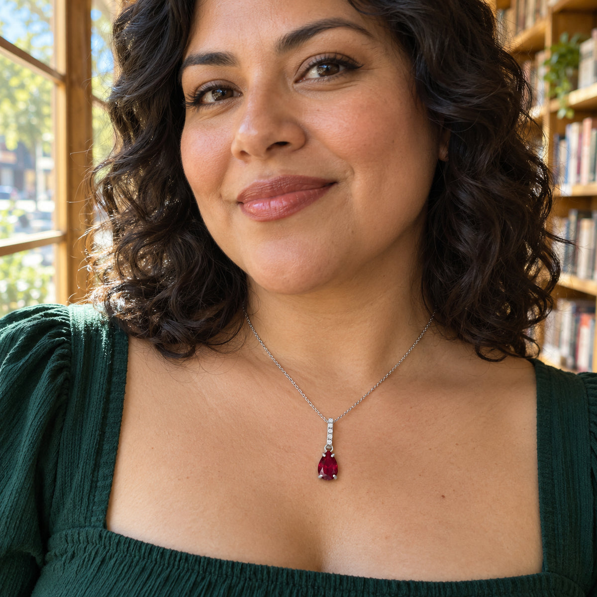 A woman with dark curls wears a silver ruby necklace above a green square-neck blouse in a bookstore.