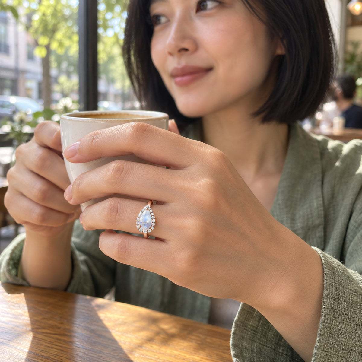 A woman with a black bob wears the rose gold moonstone and diamond ring while holding a ceramic cup in a sunlit cafe.