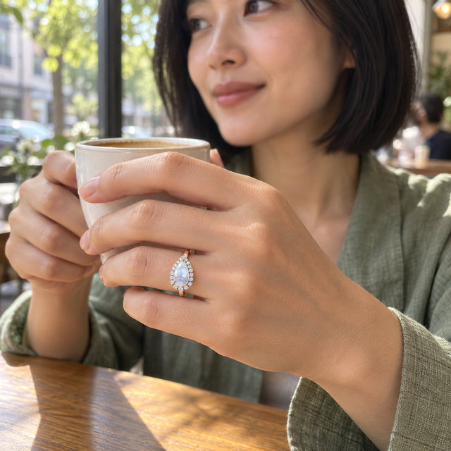 A woman with a black bob wears the rose gold moonstone and diamond ring while holding a ceramic cup in a sunlit cafe.