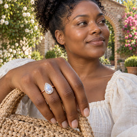 A woman with curly hair wears the rose gold moonstone and diamond ring while resting her hand on a woven straw bag in a garden.