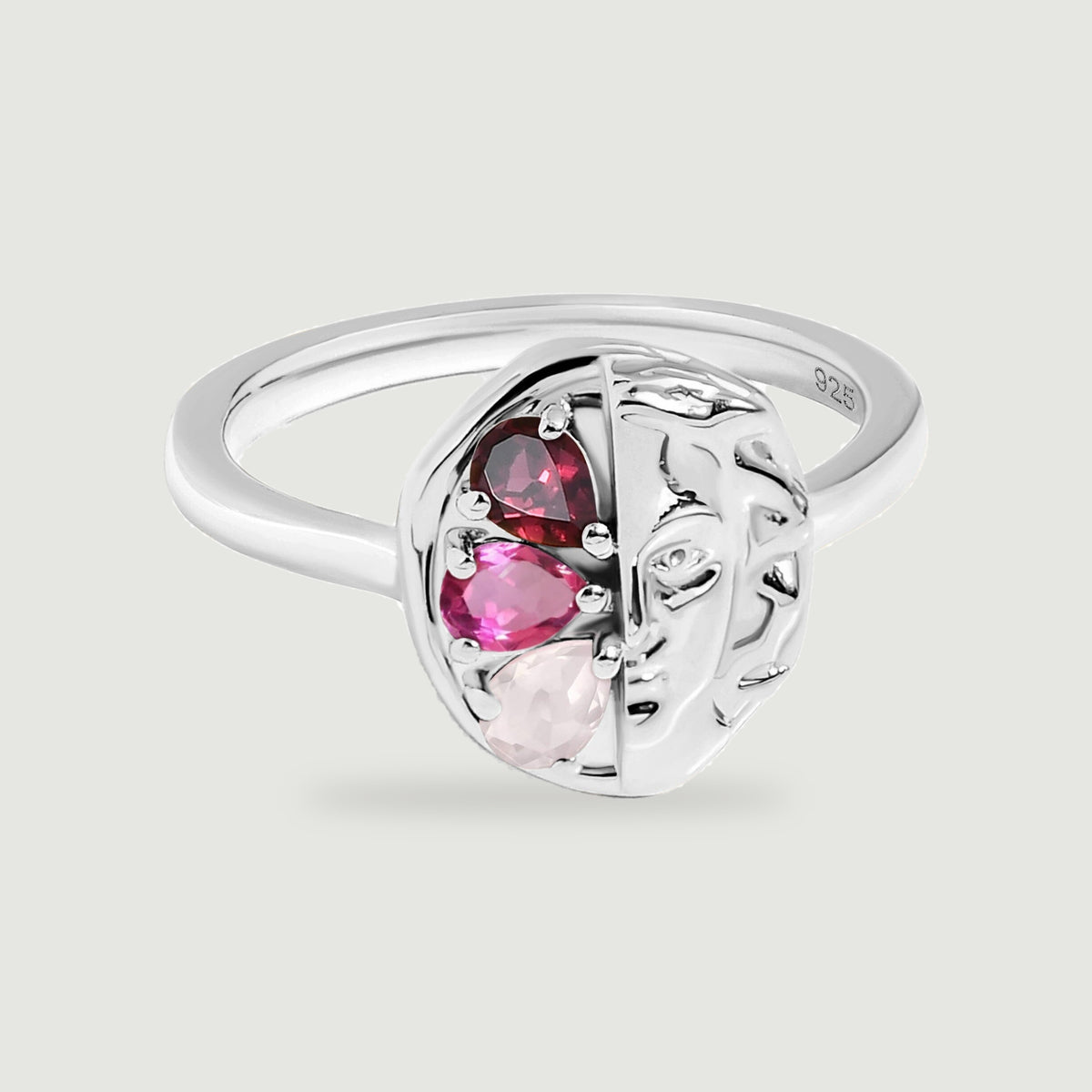 Three red and pink stones sit beside a sculpted face on a polished silver ring against a light grey background.